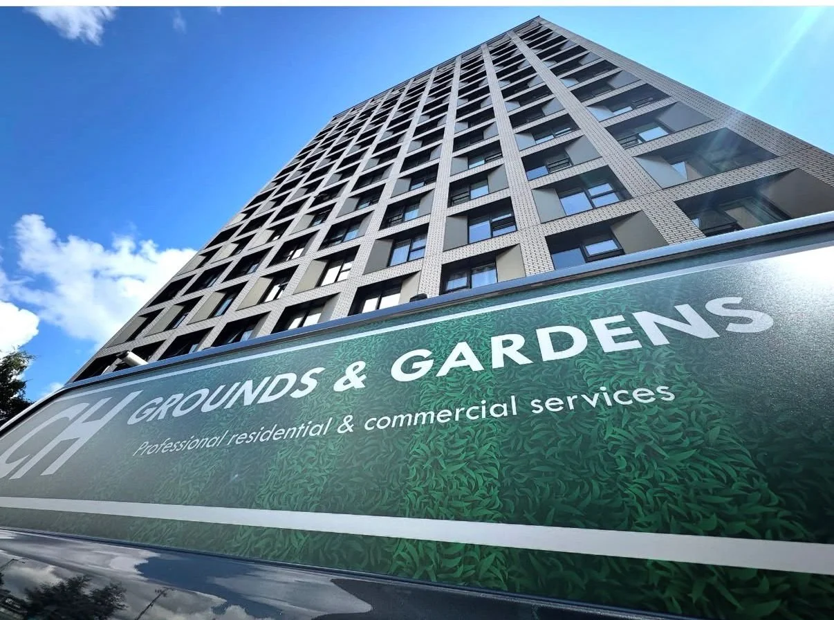 A tall building with many windows seen from a low angle, with a sign for Grounds & Gardens, a professional residential and commercial landscaping service, in the foreground.