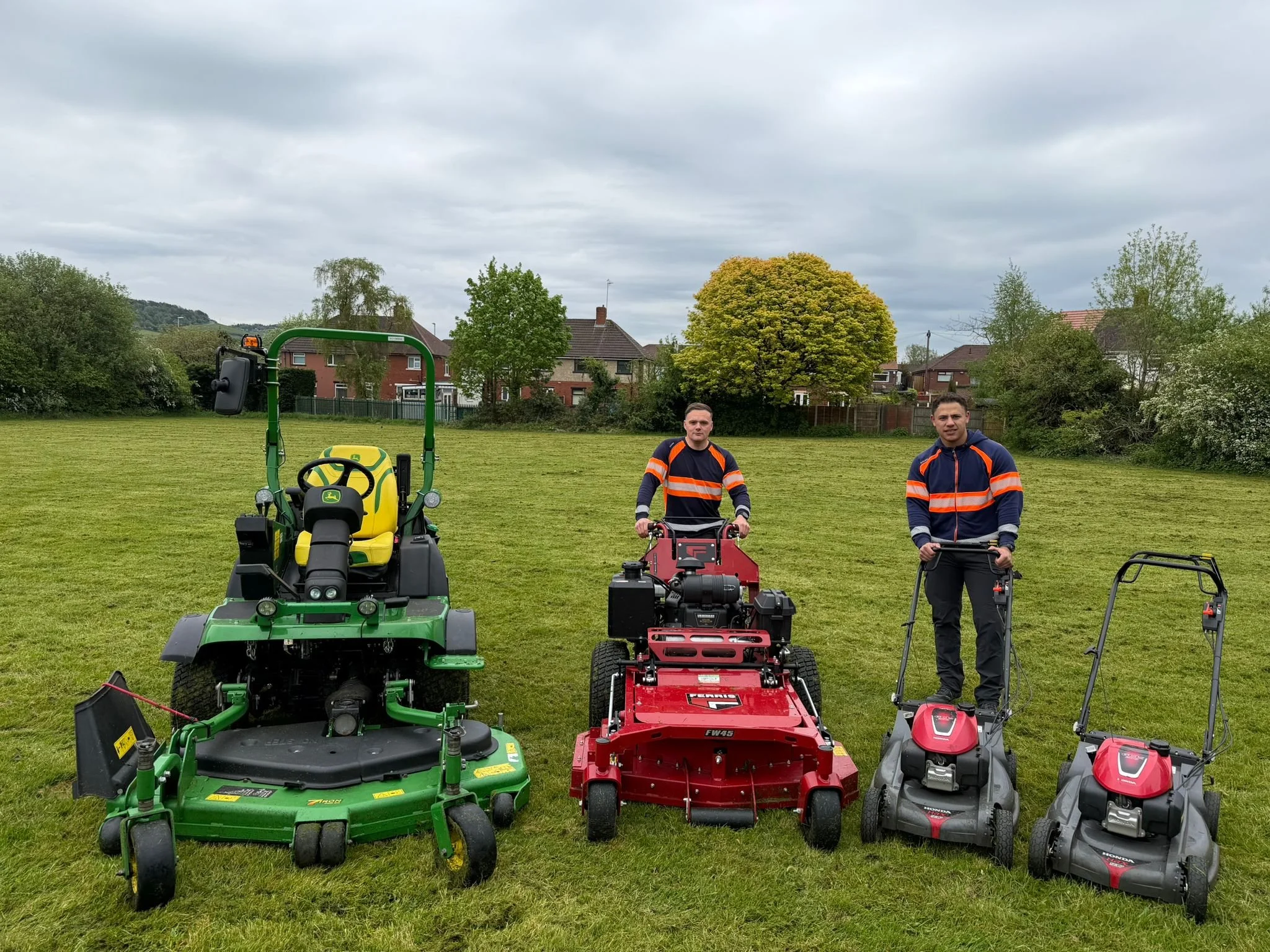 Two men standing on a grassy field surrounded by various lawn and garden equipment, including a green mower, a red mower, and a gray mower, under a cloudy sky.