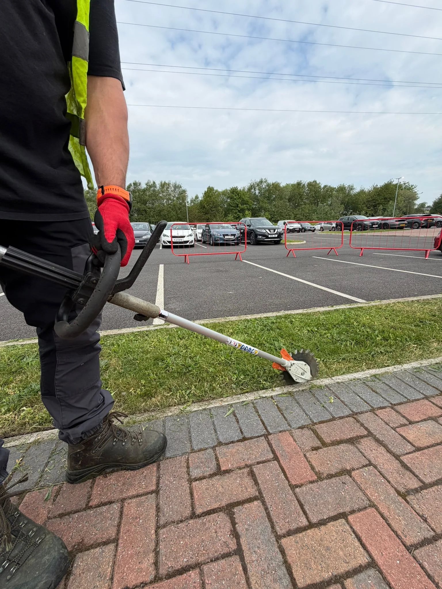 A worker wearing black pants, a black shirt, red gloves, and black boots using a grass trimmer to cut grass along the edge of a parking lot with parked cars, red safety barriers, and trees in the background.