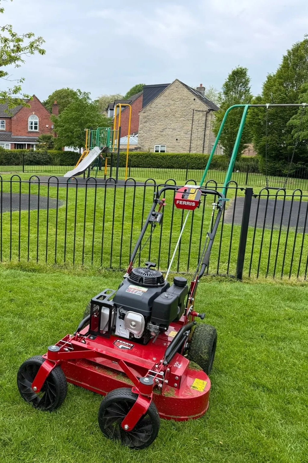 Lawn mower on a grass area in a park with a children's slide and swing set in the background, a fence surrounds the park, with houses and trees beyond.