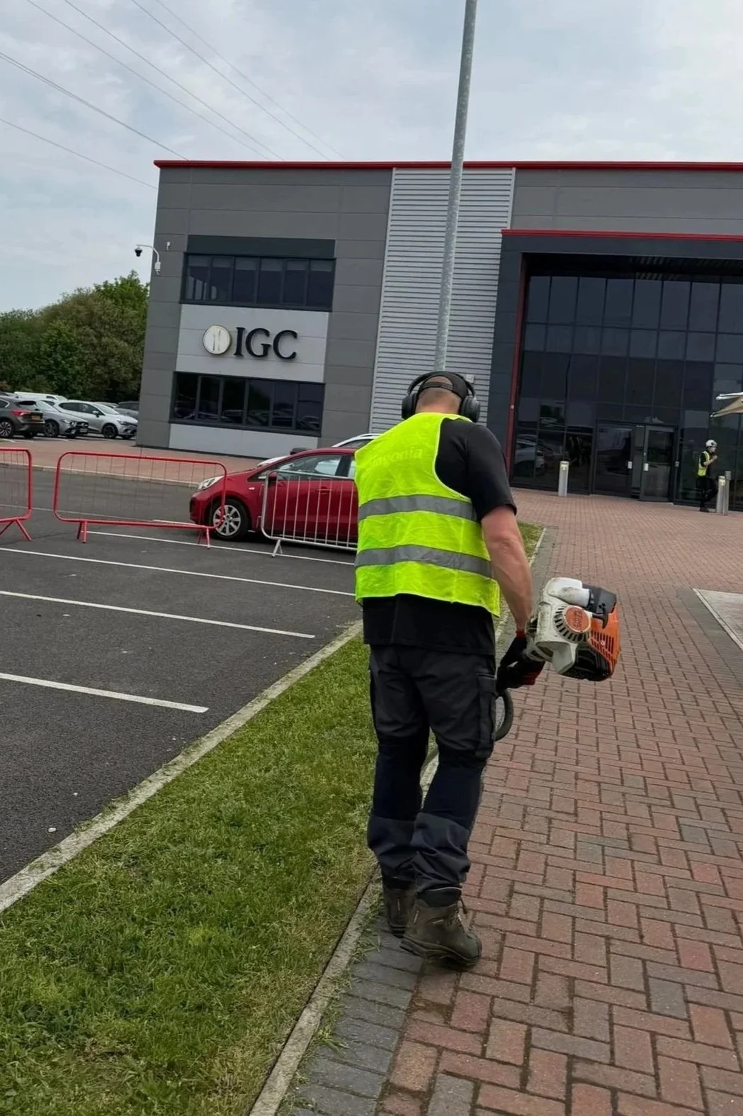 A worker wearing a yellow safety vest and black clothing is holding a leaf blower on a brick sidewalk outside a modern office building with the logo 'IGC'. There are parked cars and a gray sky in the background.