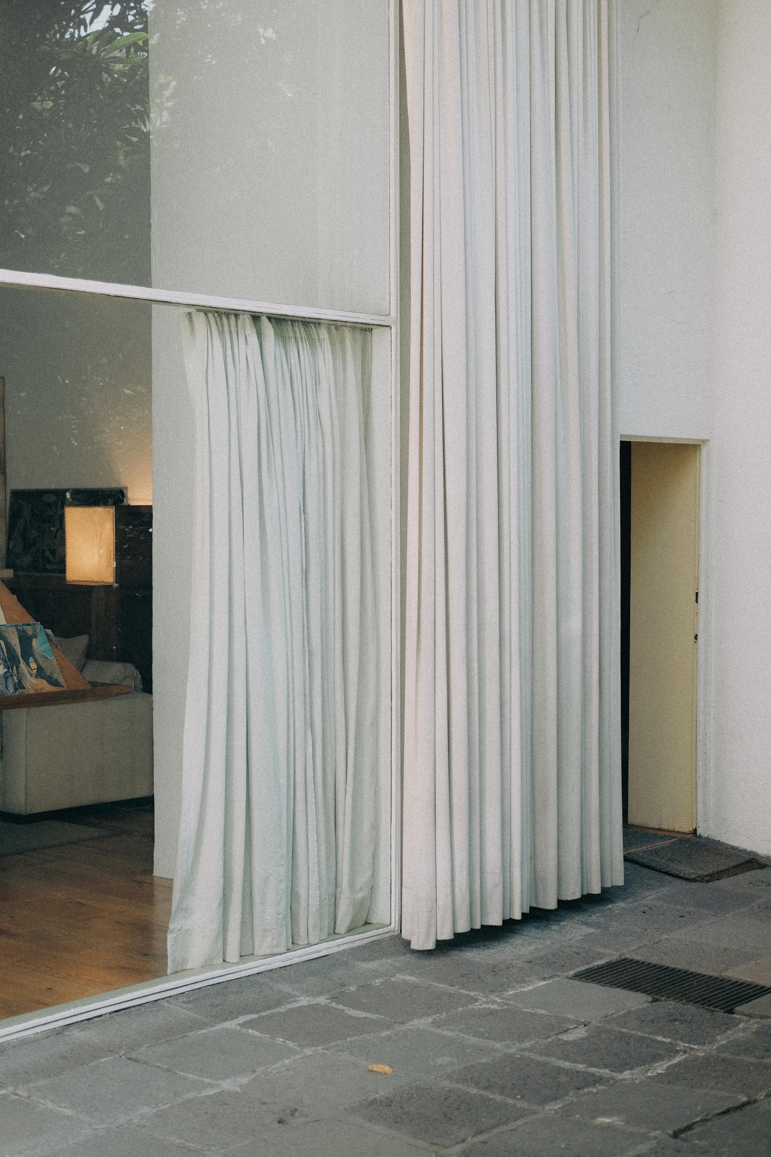 View of an interior room seen through a large glass window with white curtains, showing a sofa with pillows and a wooden lamp, with an entry door and a stone floor outside.