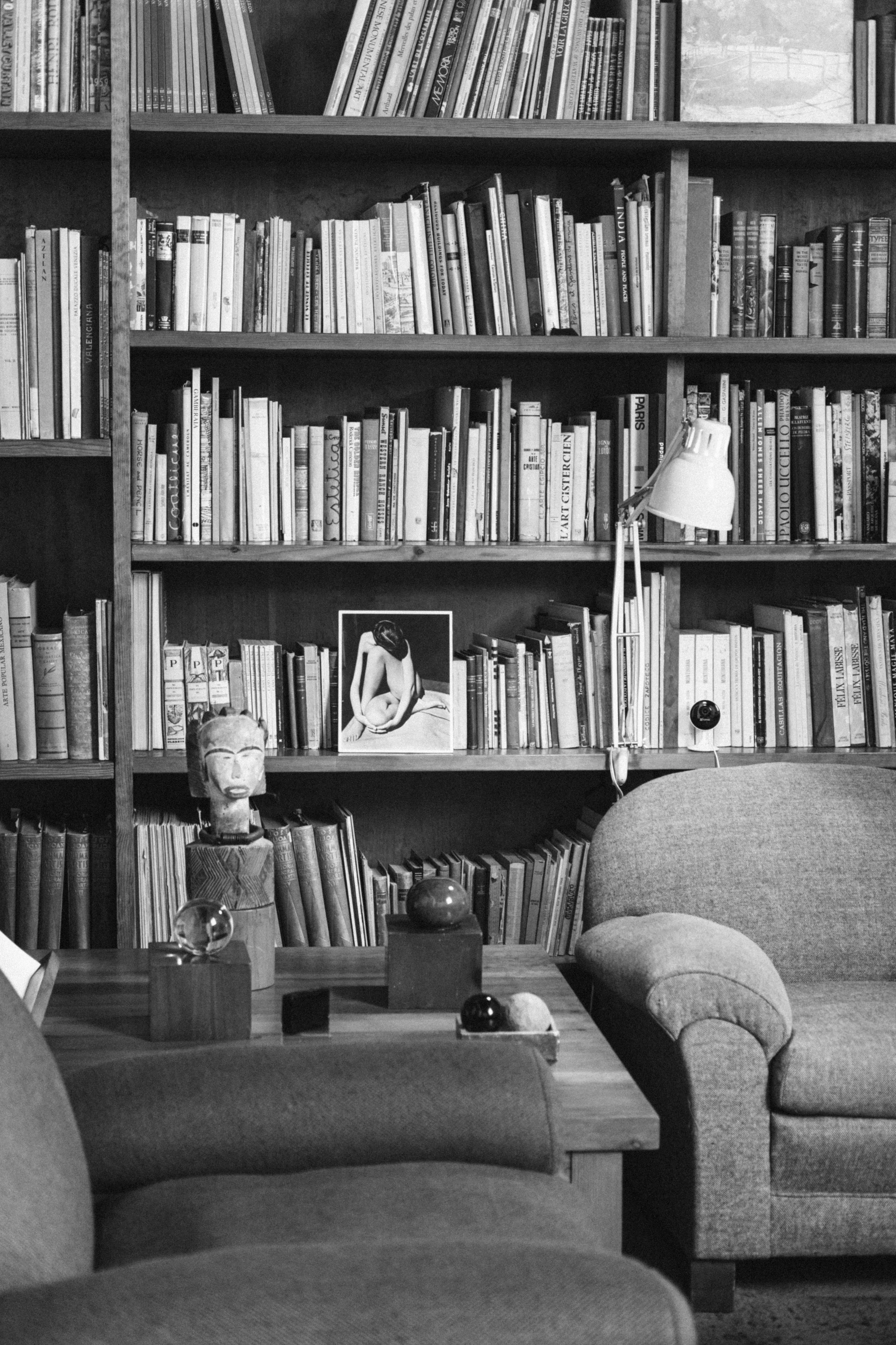 A black-and-white image of a cozy reading nook with a bookshelf filled with books, a framed photograph of a woman, and decorative objects including a statue bust and spherical ornaments on a wooden table, with comfortable armchairs nearby.