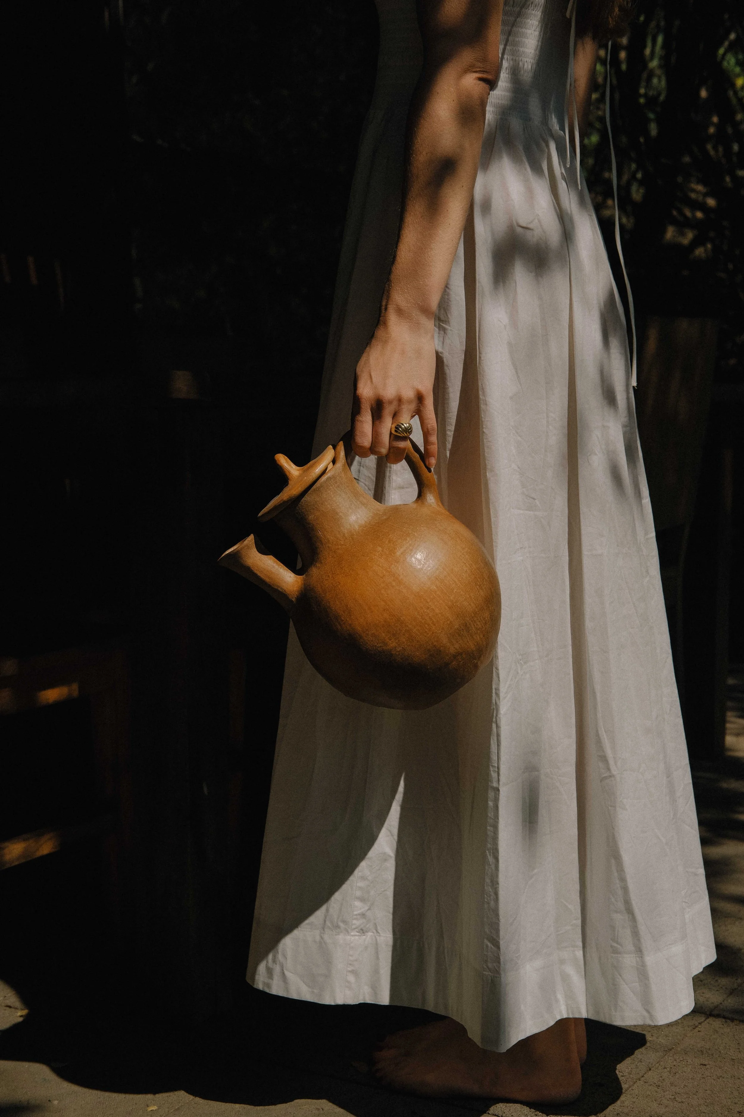 A person dressed in a long white dress is holding a large, round, brown ceramic teapot in their right hand. The photo is taken outdoors with shadows cast on the person and the ground.
