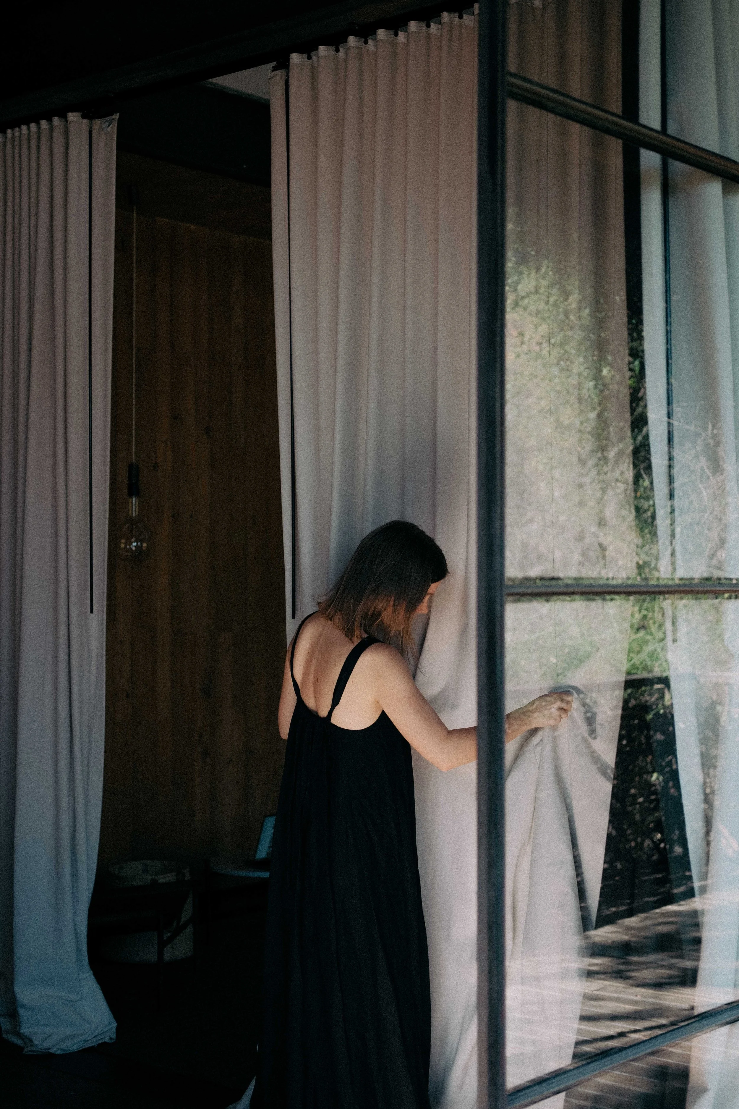 A woman in a black dress adjusting a curtain inside a room with large glass windows and white curtains.