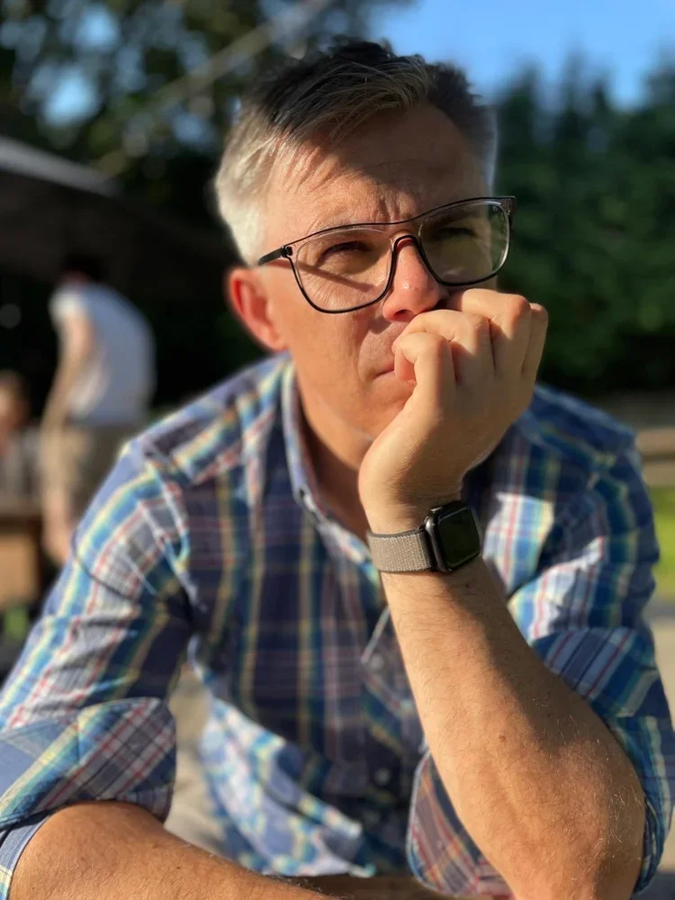 Laurie Peters with glasses resting his chin on his hand outdoors during daytime, wearing a plaid shirt and a smartwatch, with blurry background of trees and another person.