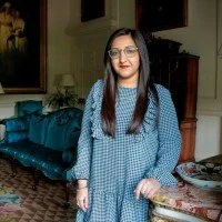 Young woman with long dark hair and glasses standing in a living room, wearing a blue patterned dress.