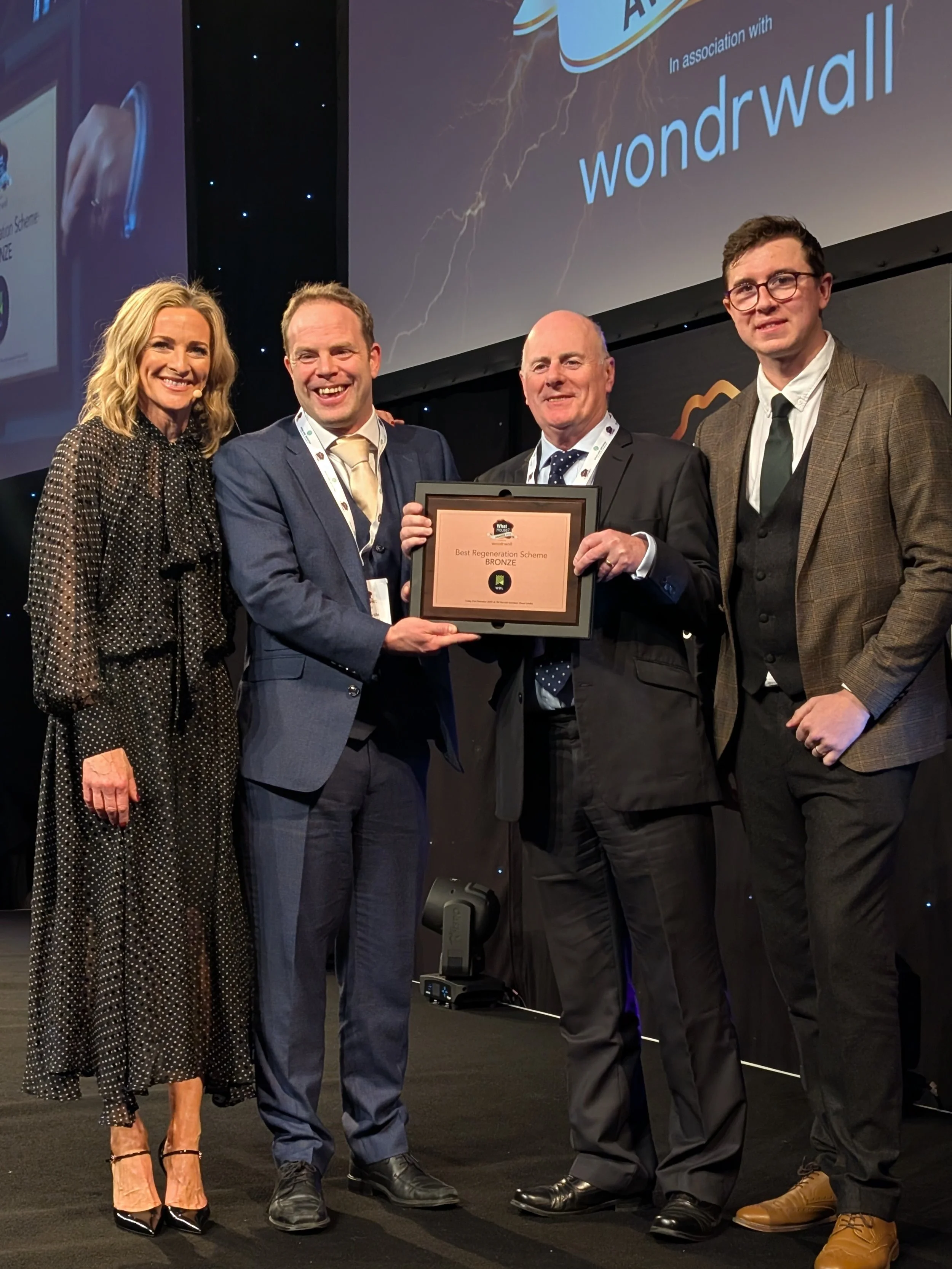 Four people standing on stage at an award ceremony, holding a plaque that reads 'Best Regeneration Scheme, Bronze'. The background features a large screen with text and a dark backdrop with small lights.