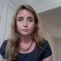 A young woman with long, wavy blonde hair wearing a dark shirt and layered necklaces, posing indoors against a neutral background.