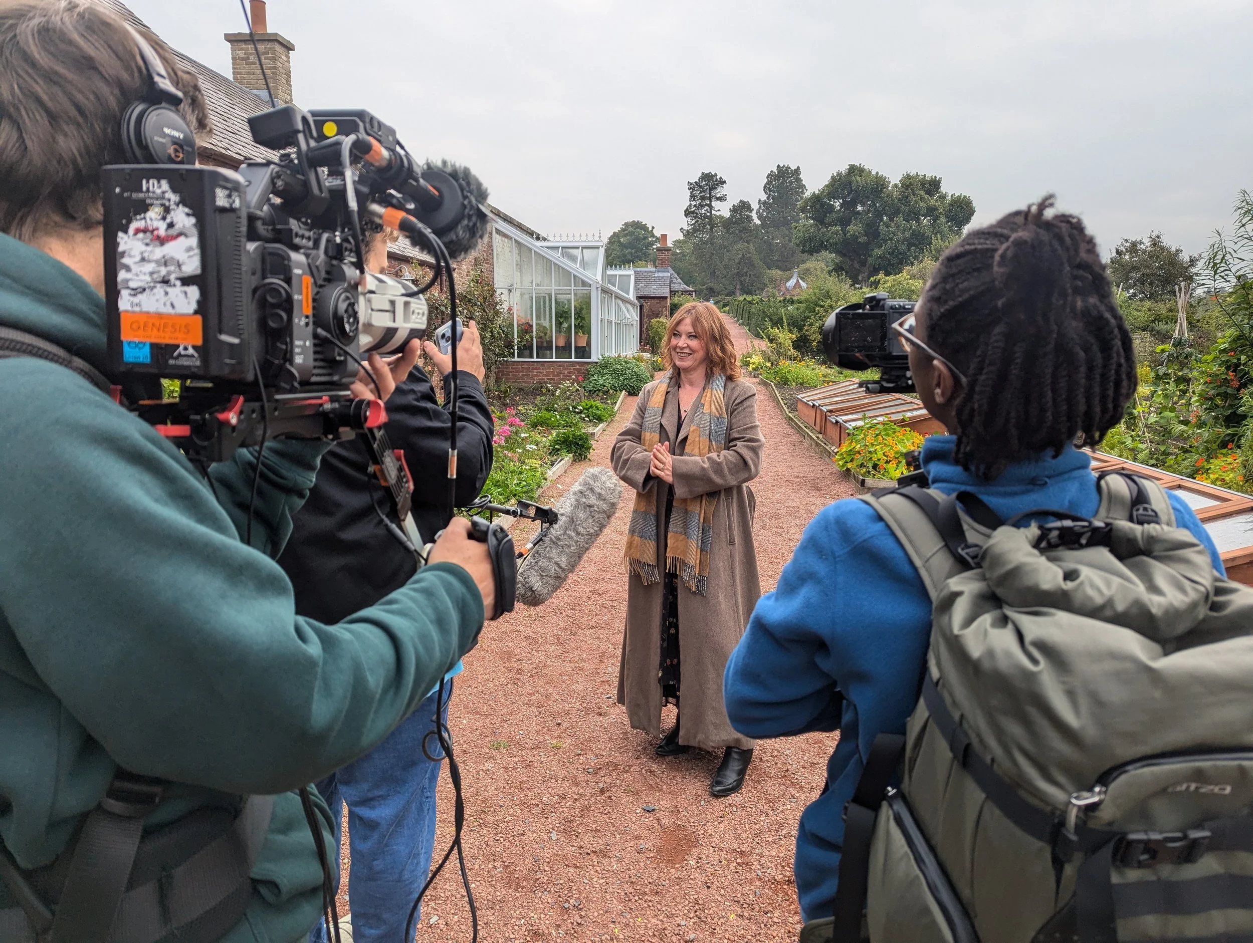 A woman with red hair, wearing a long beige coat with a multi-colored scarf, stands on a garden path being interviewed by a group of people with cameras and microphones. The garden has green plants and glass greenhouses in the background under a cloudy sky.