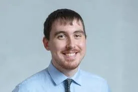 Portrait of a young man with short dark hair and a beard, smiling, wearing a light blue shirt and tie against a light blue background.