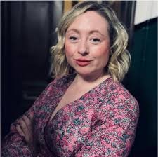 A woman with blonde, wavy hair wearing a pink patterned top, smiling slightly with a neutral background.