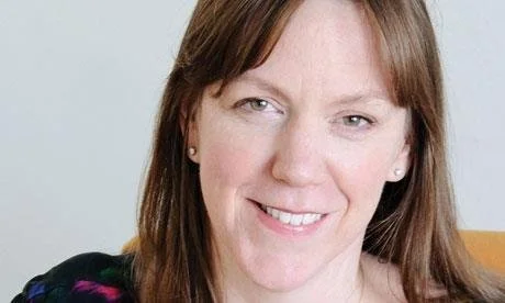 A woman with shoulder-length brown hair, smiling, wearing earrings, and a black top with colorful patterns.