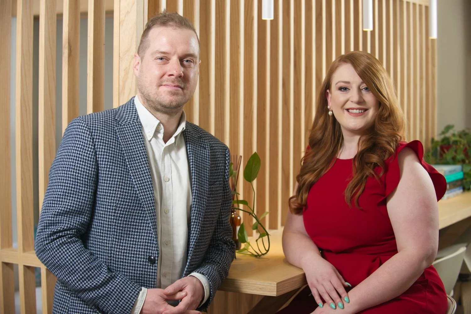 A man and a woman standing side by side indoors, smiling at the camera. The man has short hair and wears a checkered blazer over a white shirt. The woman has long curly hair and wears a red dress with ruffled sleeves, and pearl earrings. They are in front of a wooden slatted wall with some green plants and books on a counter behind them.
