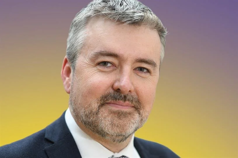 A middle-aged man with gray hair and beard, wearing a suit and tie, smiling softly at the camera with a colorful gradient background.
