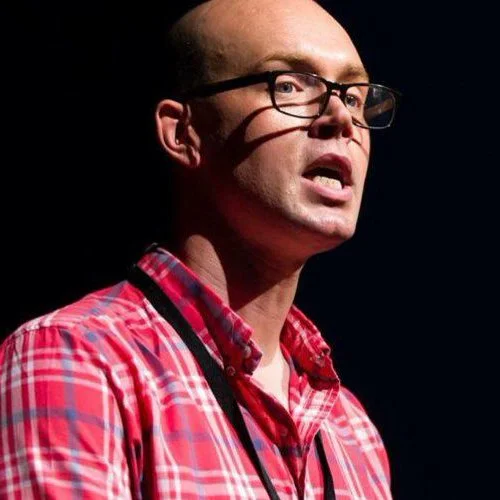 A man with glasses and short hair wearing a red plaid shirt speaking or presenting.