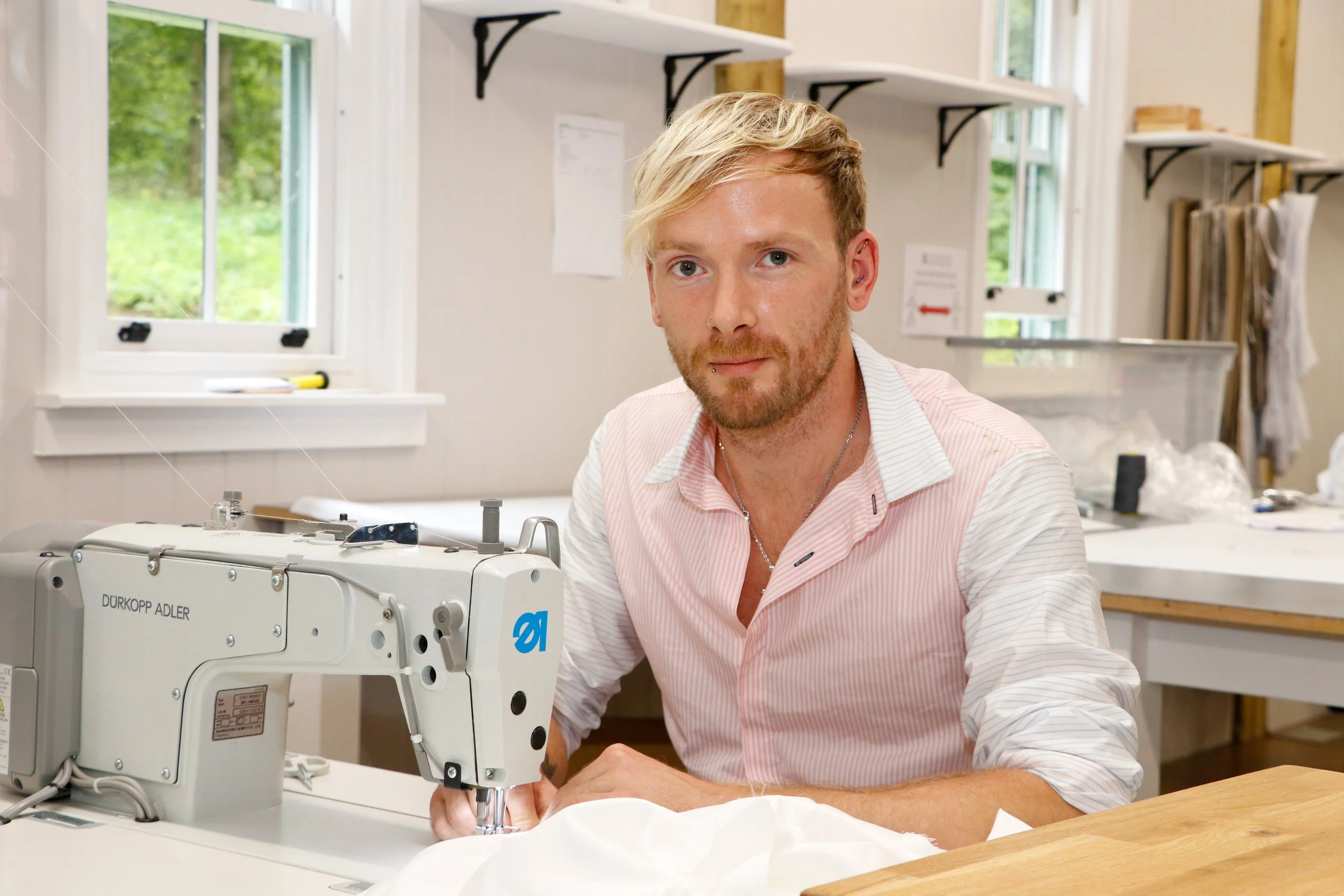 A man with blond hair and a beard working with a sewing machine in a bright room with windows and shelves.