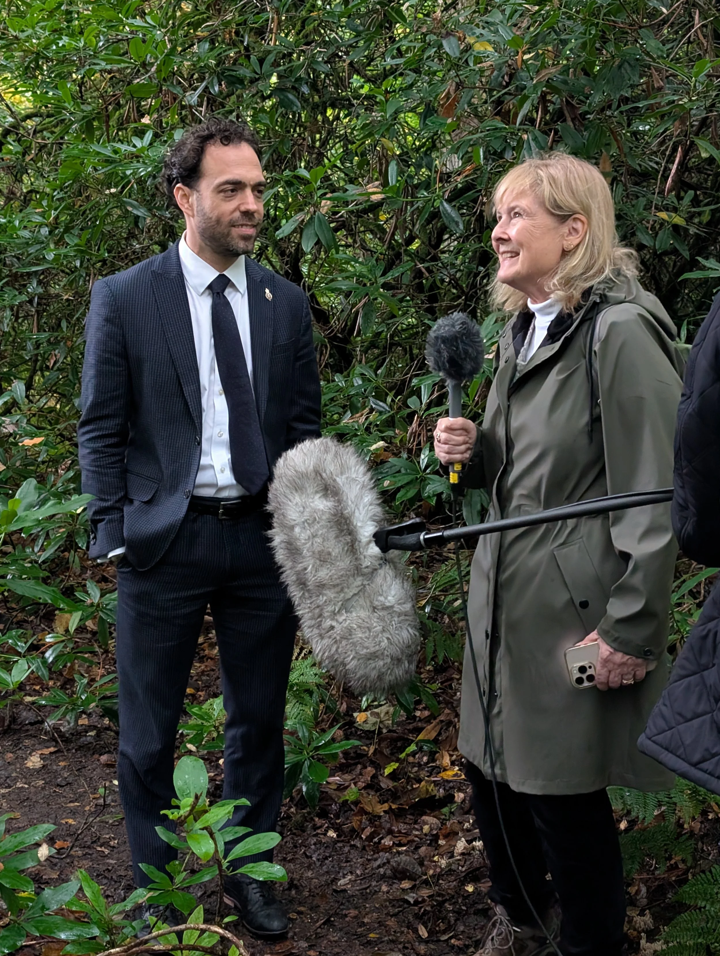 A woman interviews a man with a microphone and recording equipment in a dense, green forest.