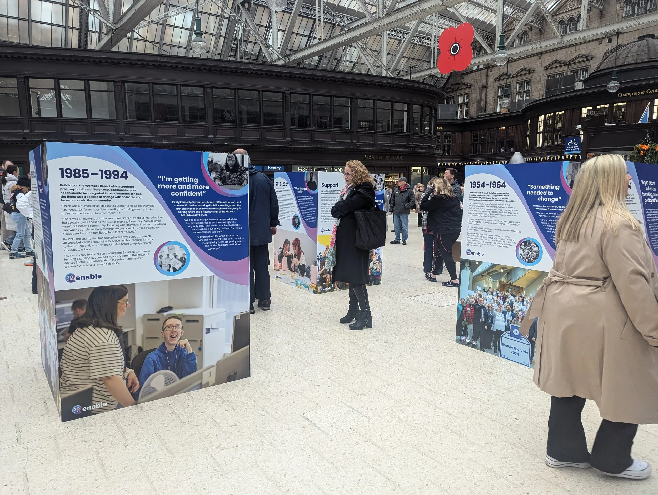 Exhibit panels inside a historic building with high glass ceiling, displaying information about community support, with people viewing and reading the displays.