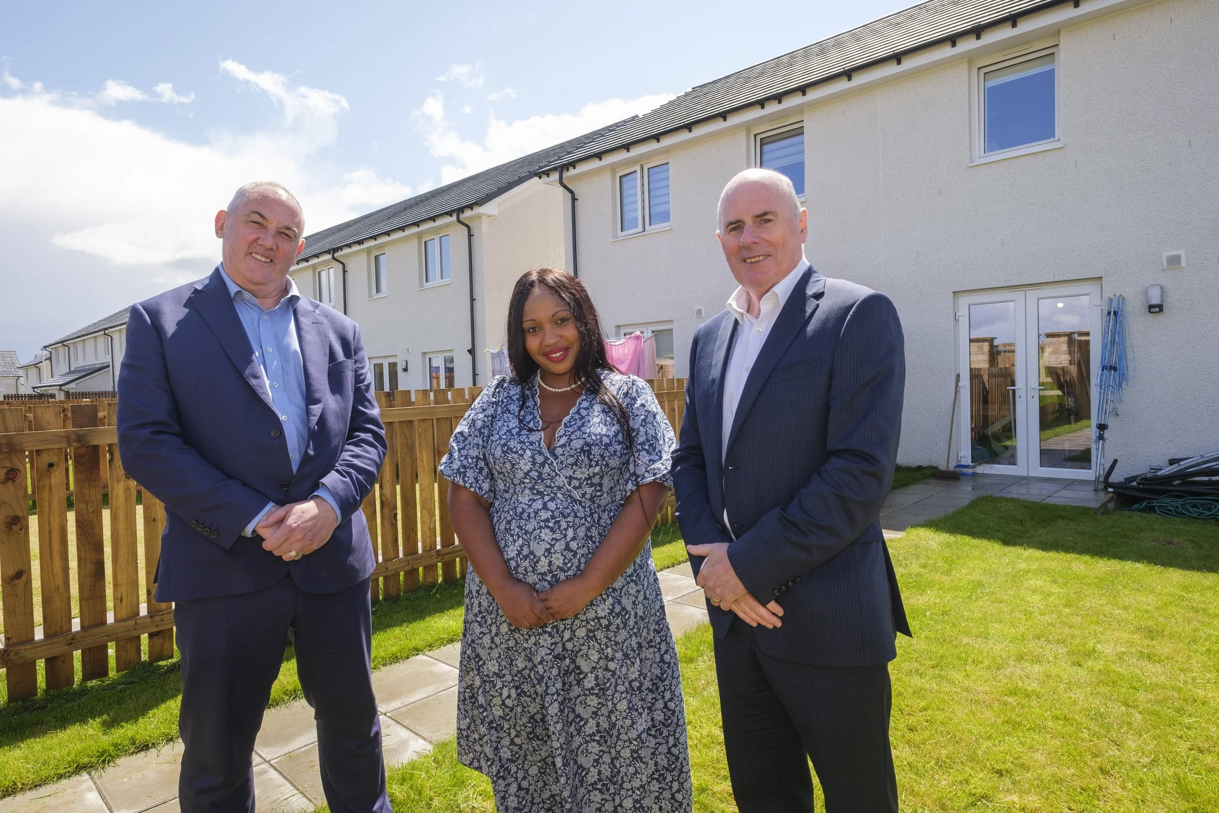 Three people standing outside in a backyard with a row of multi-story houses in the background on a sunny day.