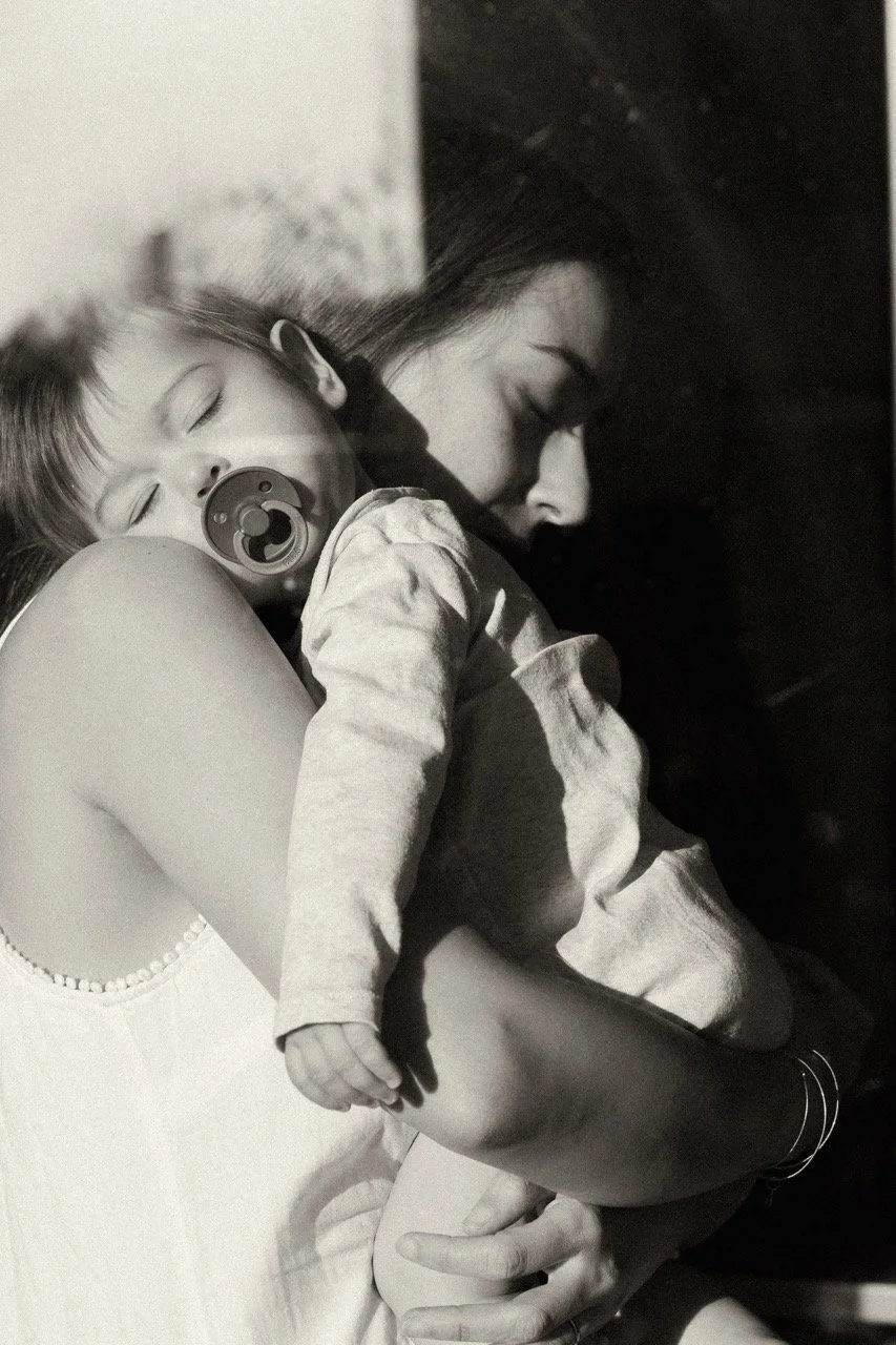Intimate black and white portrait of a mother holding her sleepy baby close — a quiet moment of exhaustion and love held together. Motherhood photography by Noa Nguyen, Tokyo.