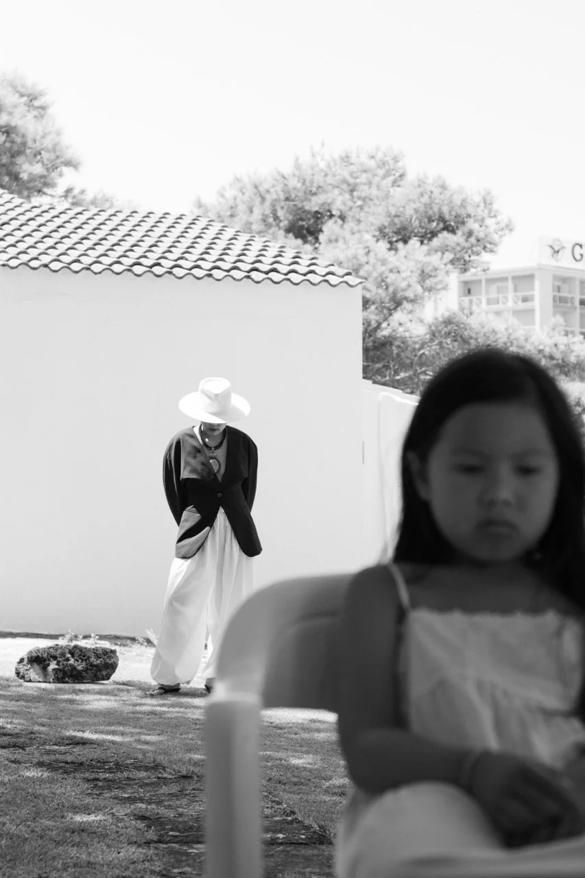 A child blurred in the foreground while a mother walks alone in the background — the distance and closeness of family life captured in one frame. Documentary family photography by Noa Nguyen.