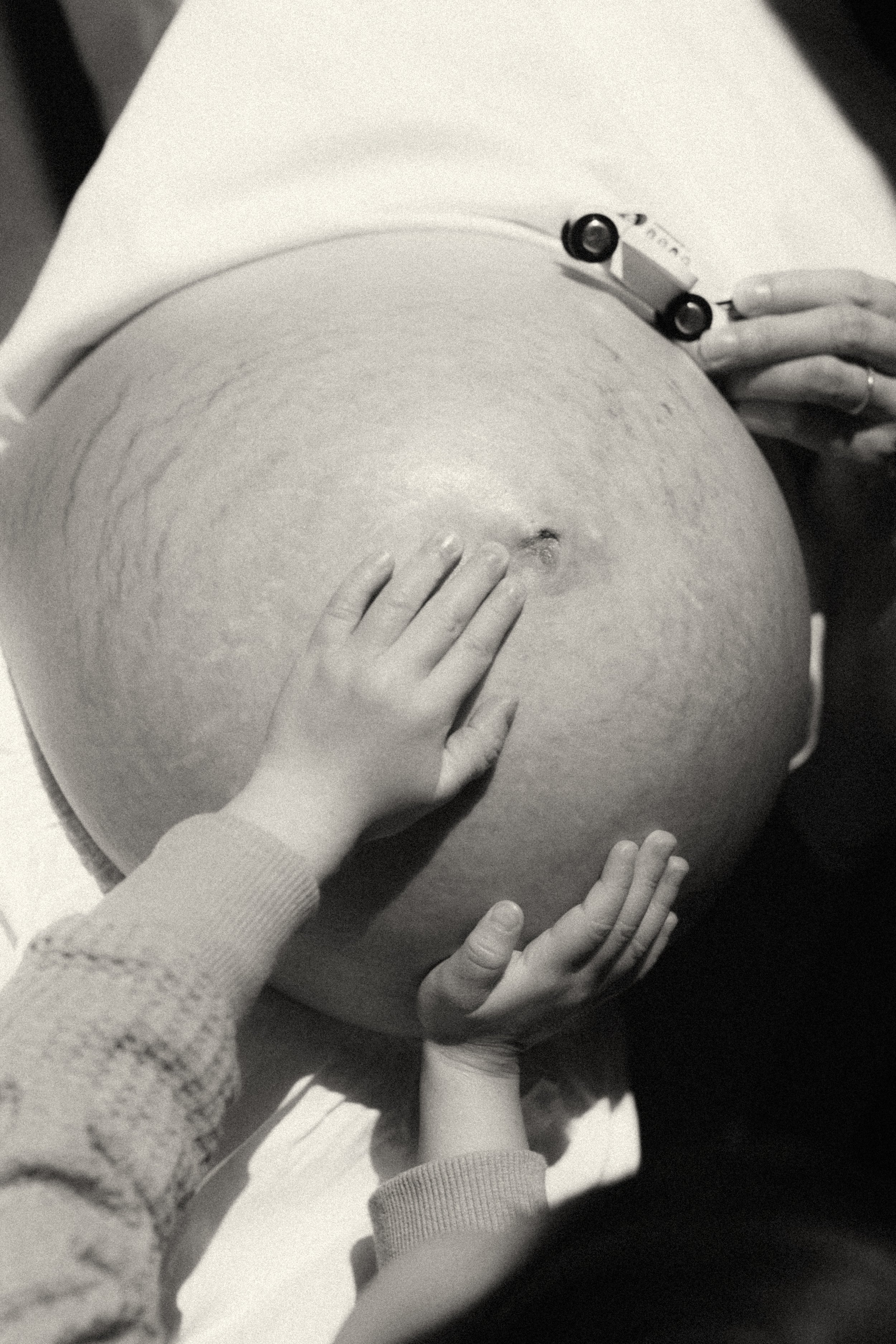 Intimate black and white maternity photograph of a toddler's small hands touching their mother's pregnant belly — the tender moment of a sibling transition, a family quietly growing into a new shape. Maternity photography by Noa Nguyen, Tokyo.
