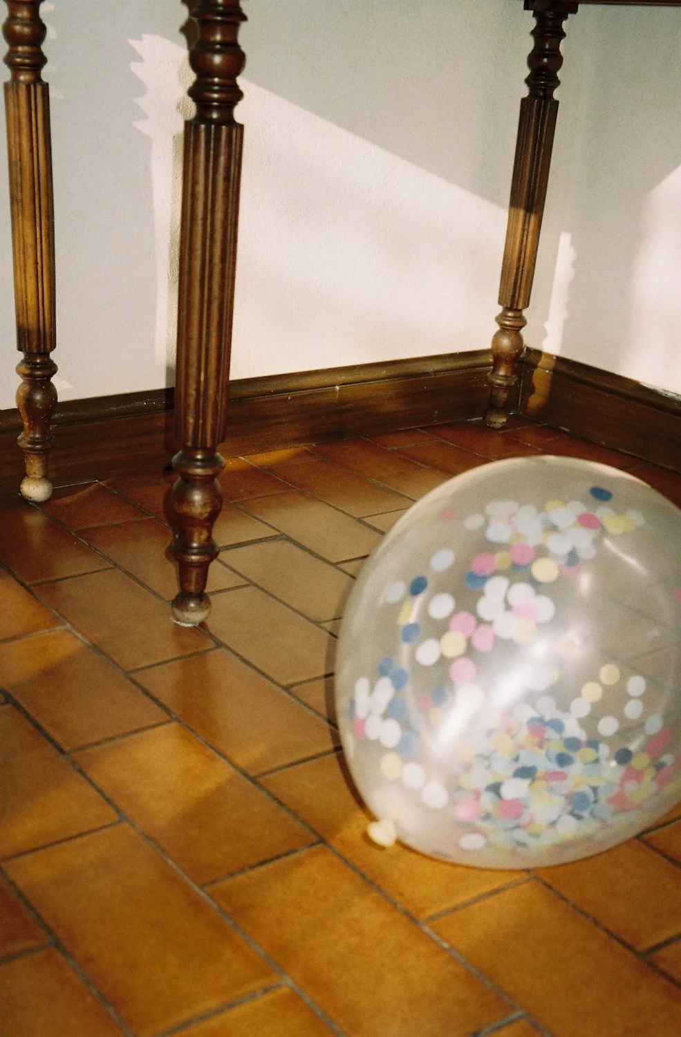 A confetti-filled balloon resting on a sunlit tile floor beneath an antique table — a quiet remnant of a family celebration, capturing the warmth of tradition at home. Family lifestyle photography by Noa Nguyen.