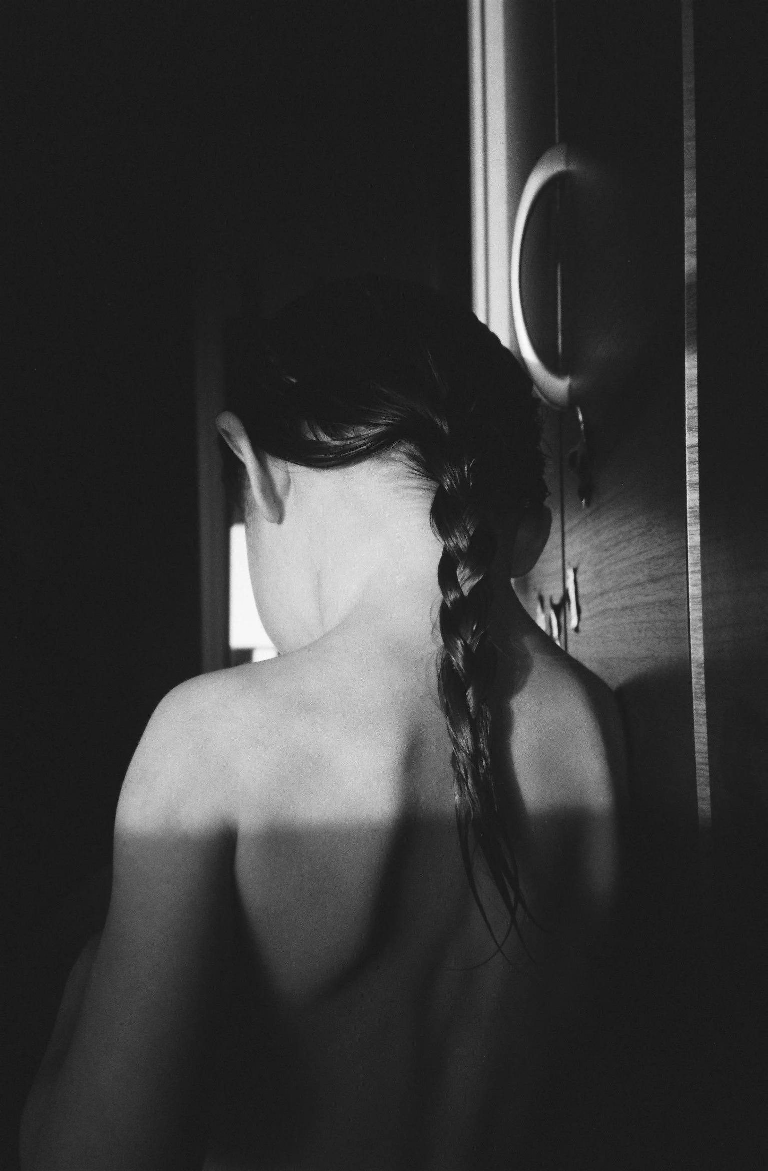 A high-contrast black and white film photograph from behind a child with a long braid, capturing the delicate detail of hair and skin in a Tokyo home.