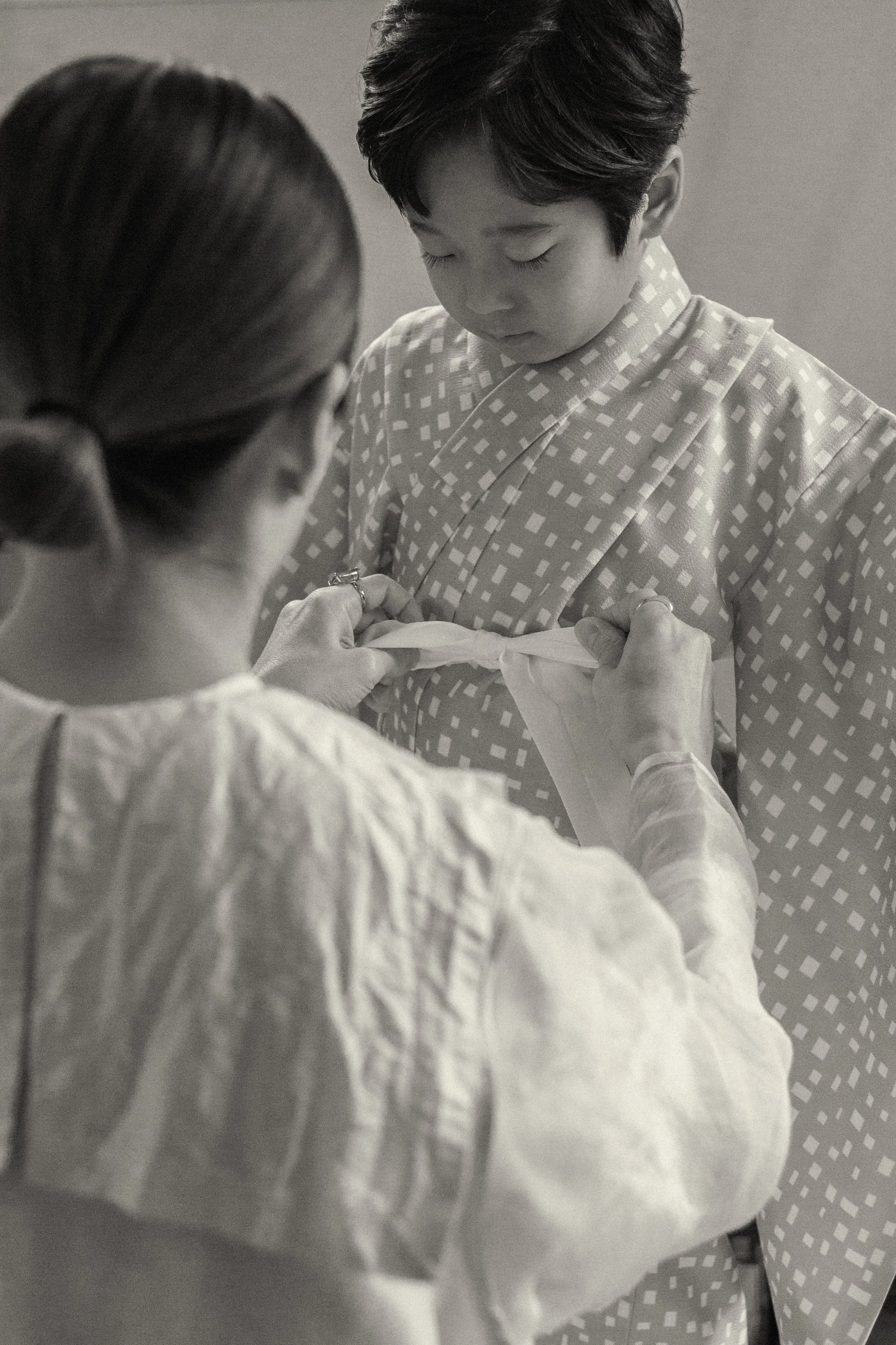 A grainy black and white photograph of a mother tying a kimono obi for her son, documenting the quiet preparation of a Shichi-Go-San celebration at home in Tokyo.