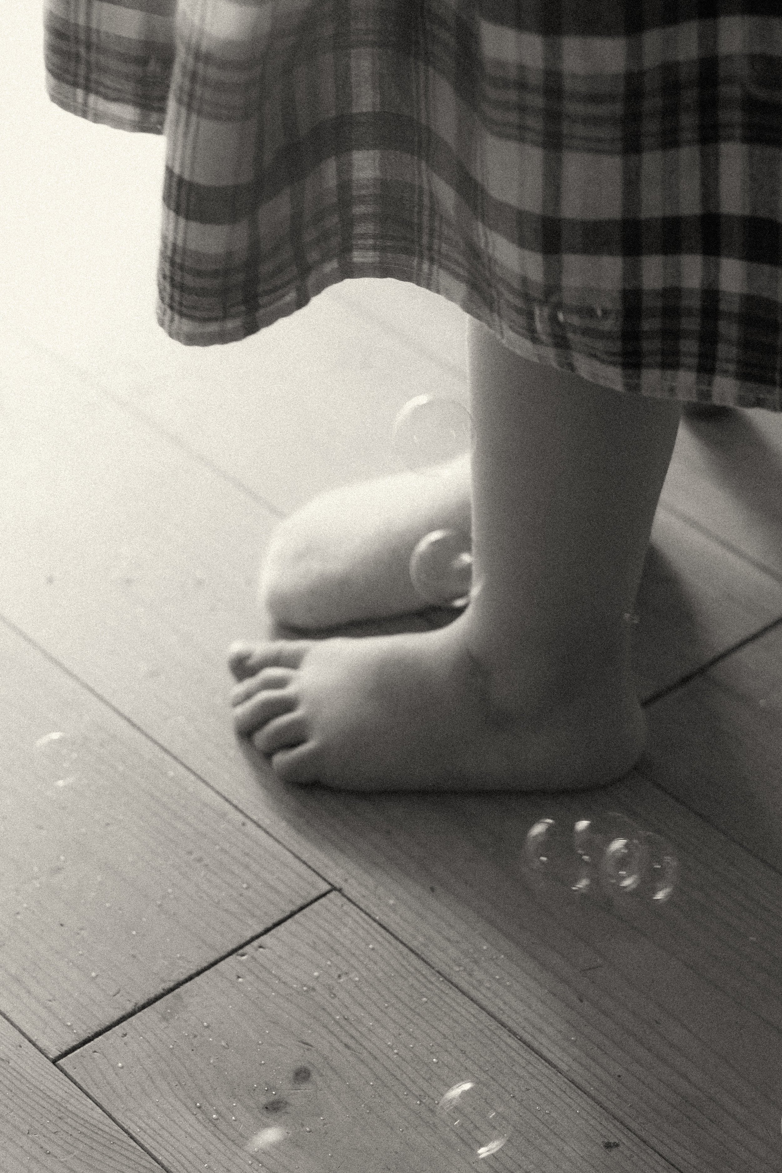 A black and white photograph close-up of a toddler's bare feet on wooden floors with soap bubbles, documenting authentic childhood play.
