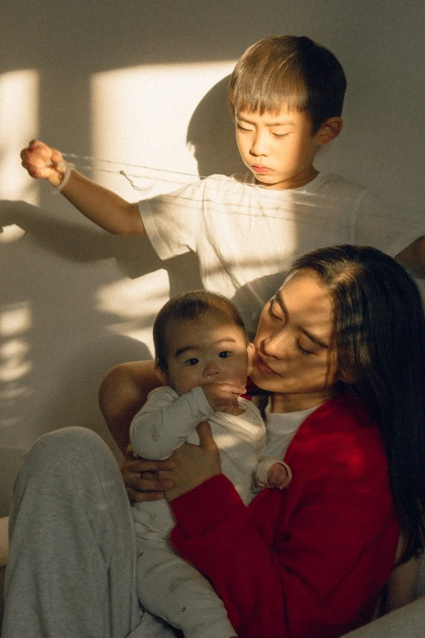 A mother holds her baby while her toddler plays beside her in warm morning light — an unposed everyday family moment. Family lifestyle photographer in Tokyo for expat families.