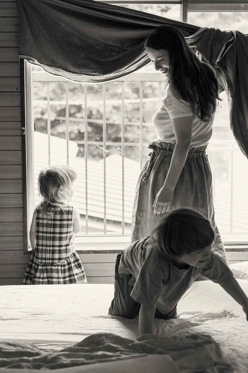 Black and white family lifestyle photograph of a mother making the bed while two young children play around her — the beautiful, chaotic energy of everyday life with toddlers, captured at home. Family lifestyle photography by Noa Nguyen.