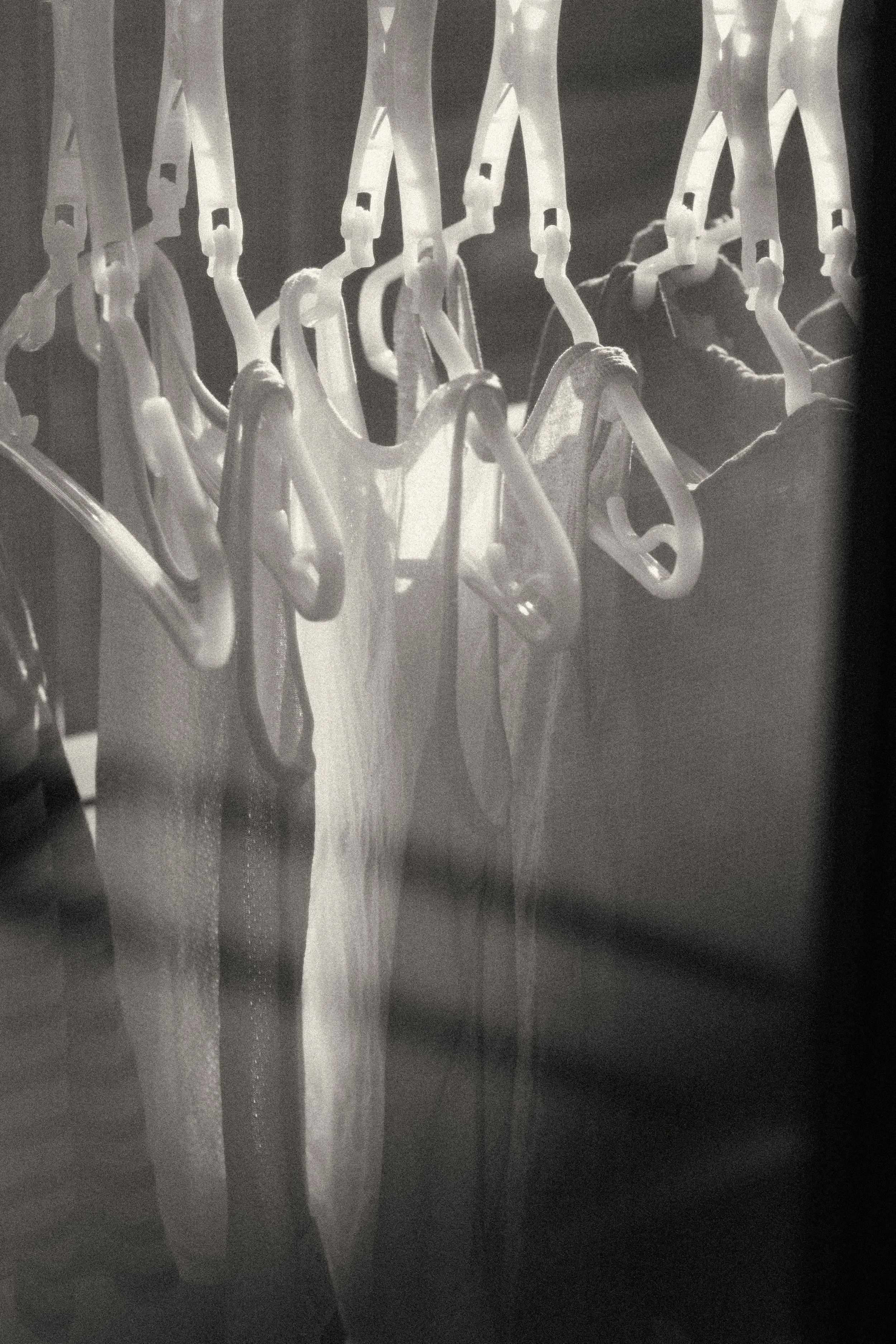 A grainy black and white photograph of clothes hanging on a rack with dramatic light and shadows, documenting the quiet beauty of daily chores, endless mother’s works.