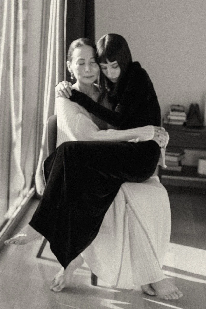 The same mother, years later — holding her grown daughter by a window. A portrait of motherhood past and present, tender love across time. editorial Family photography by Noa Nguyen.