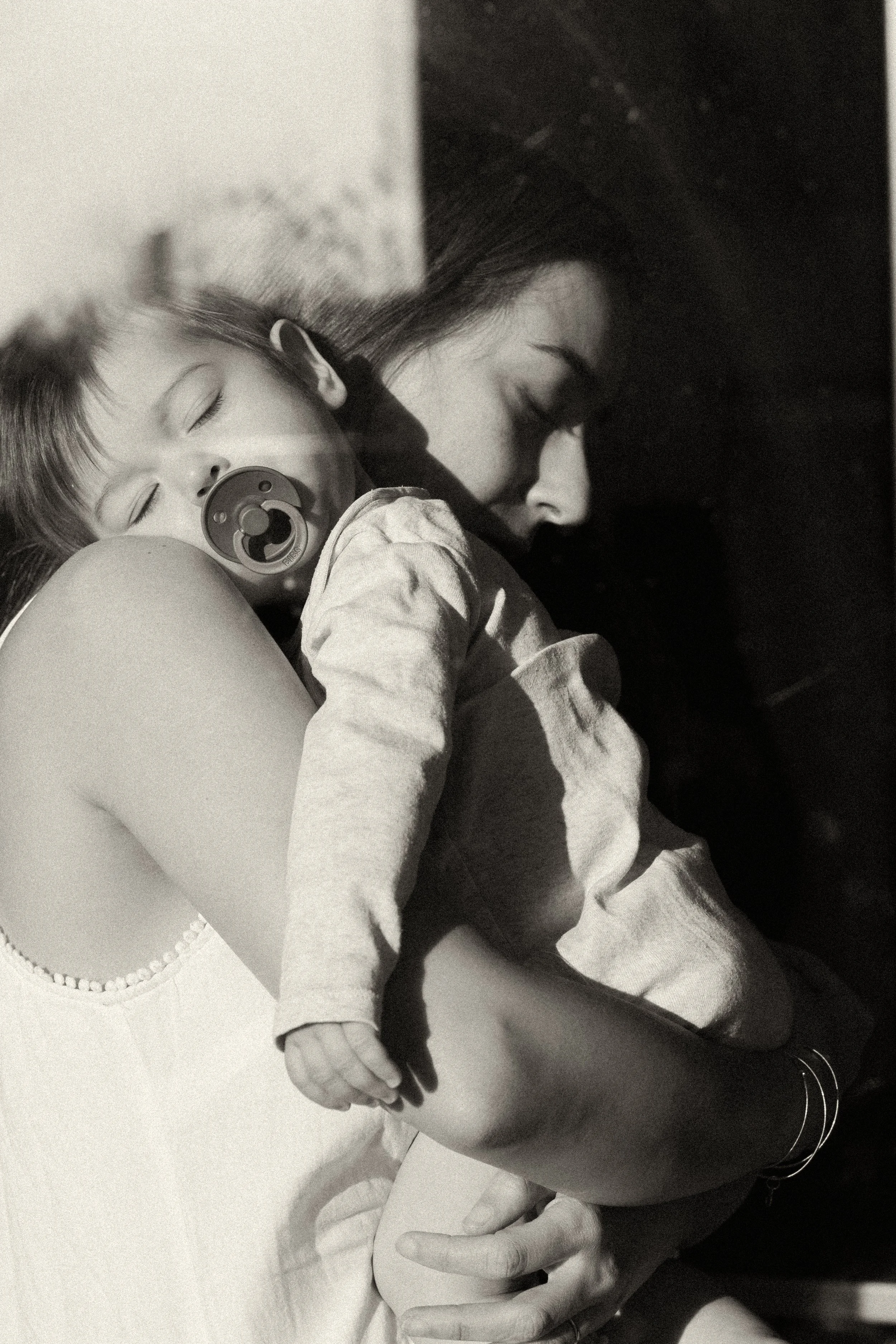 A grainy black and white portrait of a mother holding her growing child in a high-contrast embrace, documenting the quiet strength and exhaustion of daily motherhood in Paris.
