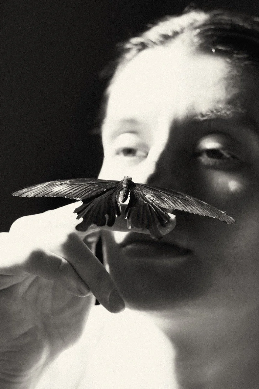 A mother holds a butterfly close to her face in dramatic light — an editorial portrait exploring transformation and identity in motherhood. Fine art photography by Noa Nguyen. Featured on Photo Vogue