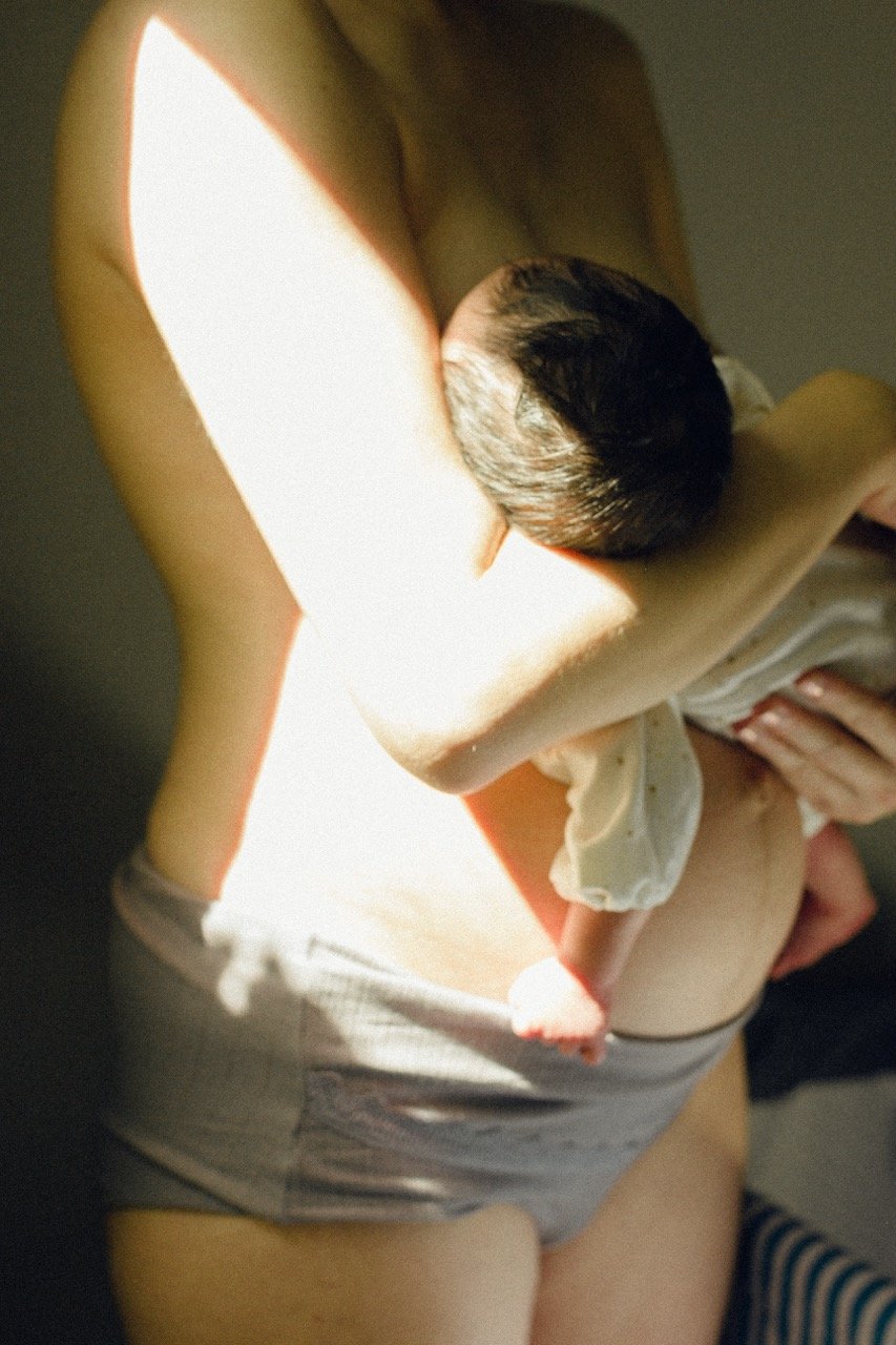 Intimate photograph of a mother holding her newborn against her bare skin in warm morning light — an honest and tender moment from the early days of motherhood. English speaking newborn photographer in Tokyo by Noa Nguyen.