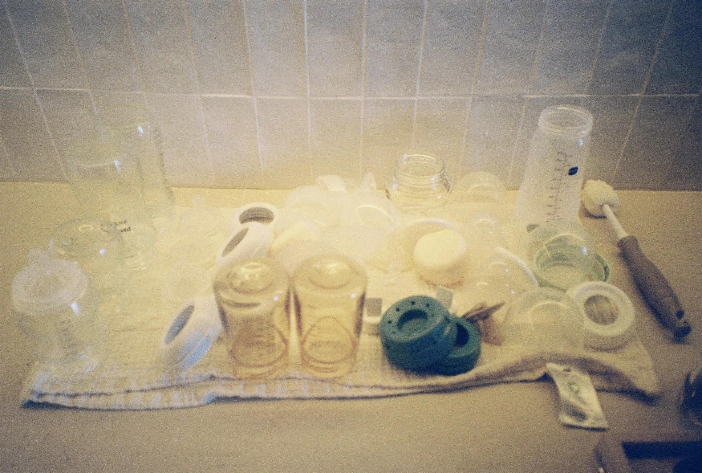 A grainy film photograph of baby bottles and milk pumping equipment drying on a kitchen counter, documenting the daily reality of motherhood in a Paris home.