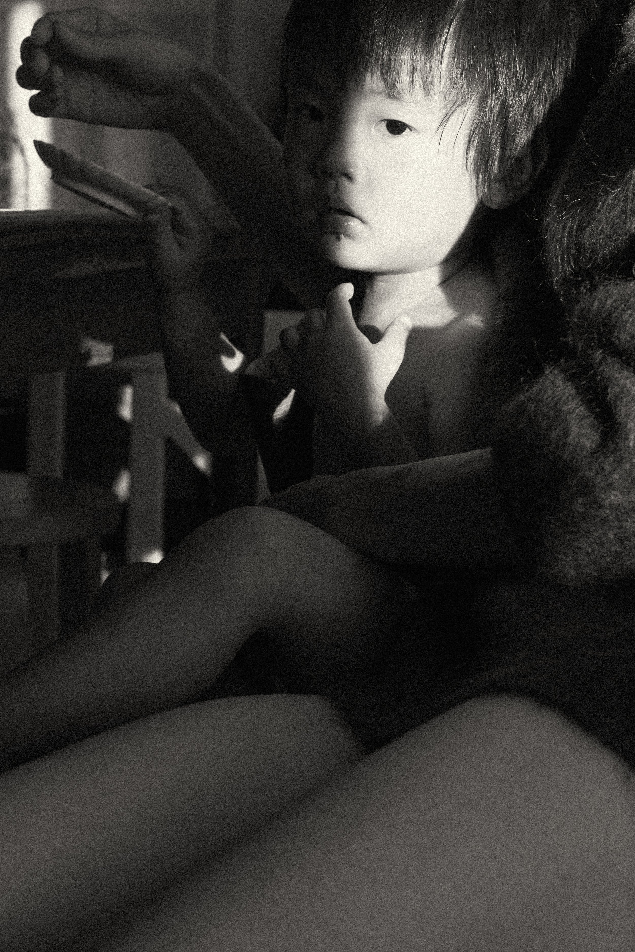 A close-up black and white portrait of a young child's face in soft natural light while sitting on their mother's lap, showcasing an unposed and intimate documentary style.