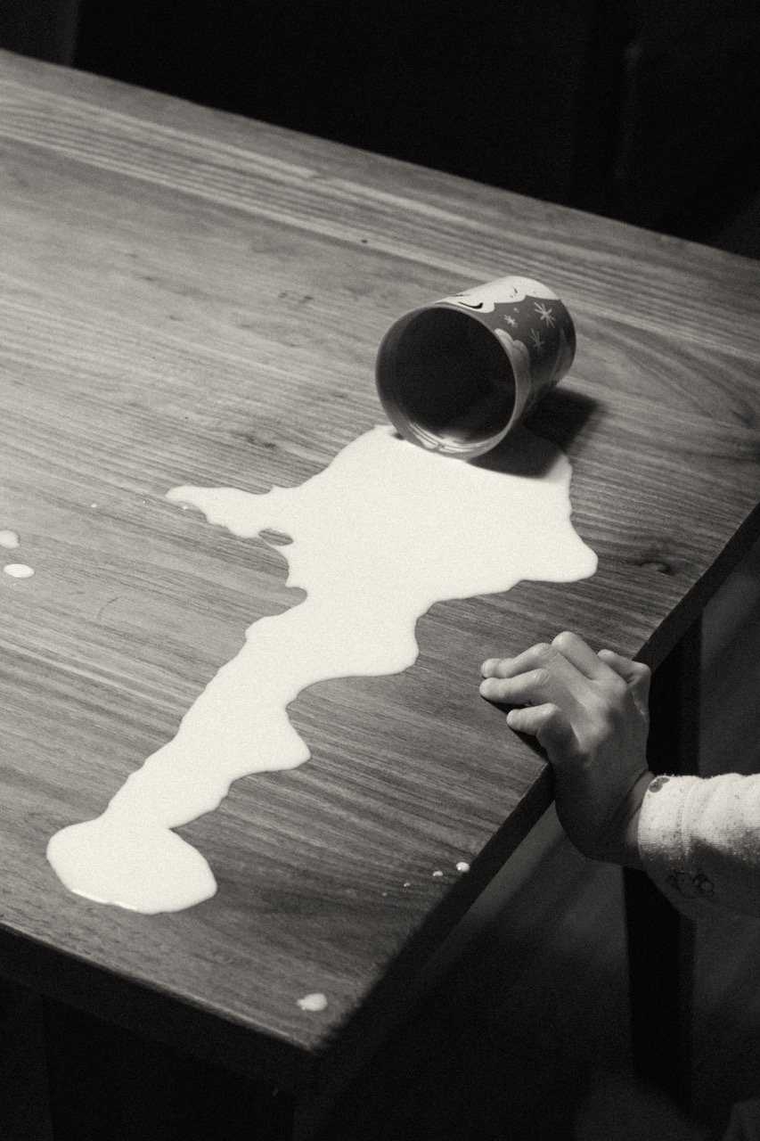 Black and white photograph of spilled milk spreading across a wooden table, a small child's hand at the edge — the beautiful, imperfect chaos of daily family life. Tokyo lifestyle family photography by Noa Nguyen.