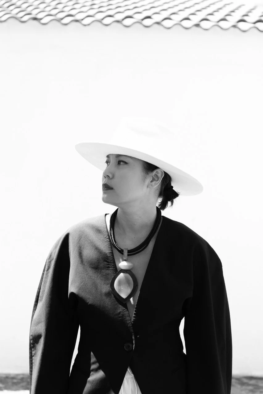 Editorial black and white portrait of a stylish mother in a wide brim hat against a white wall — a woman who remains herself through motherhood. Lifestyle portrait photography by Noa Nguyen.