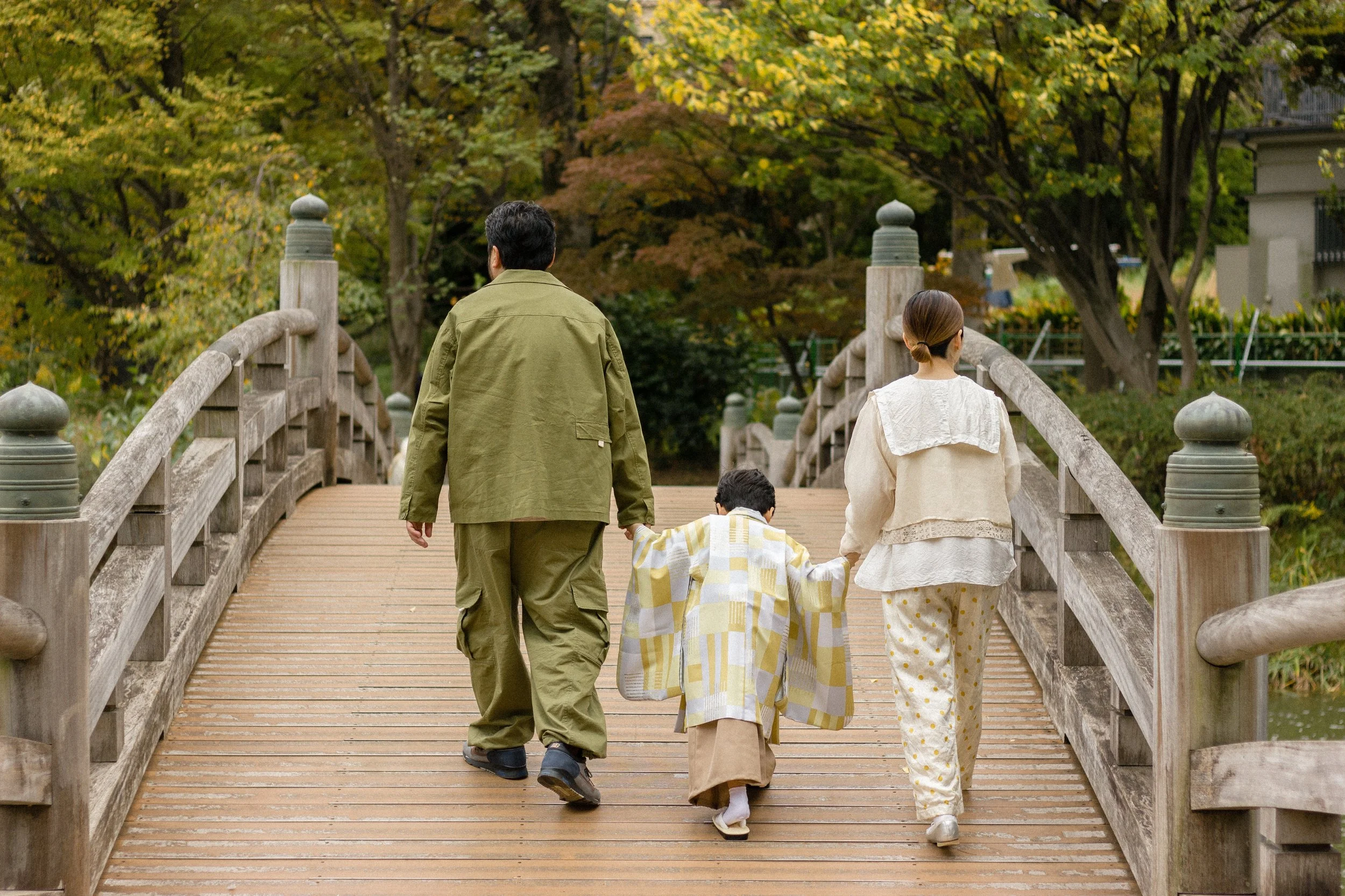 A wide-angle shot of a family walking across a wooden bridge at a Tokyo shrine during Shichi-Go-San, featuring a child in a traditional kimono and sharing an intimate moment of the family together.