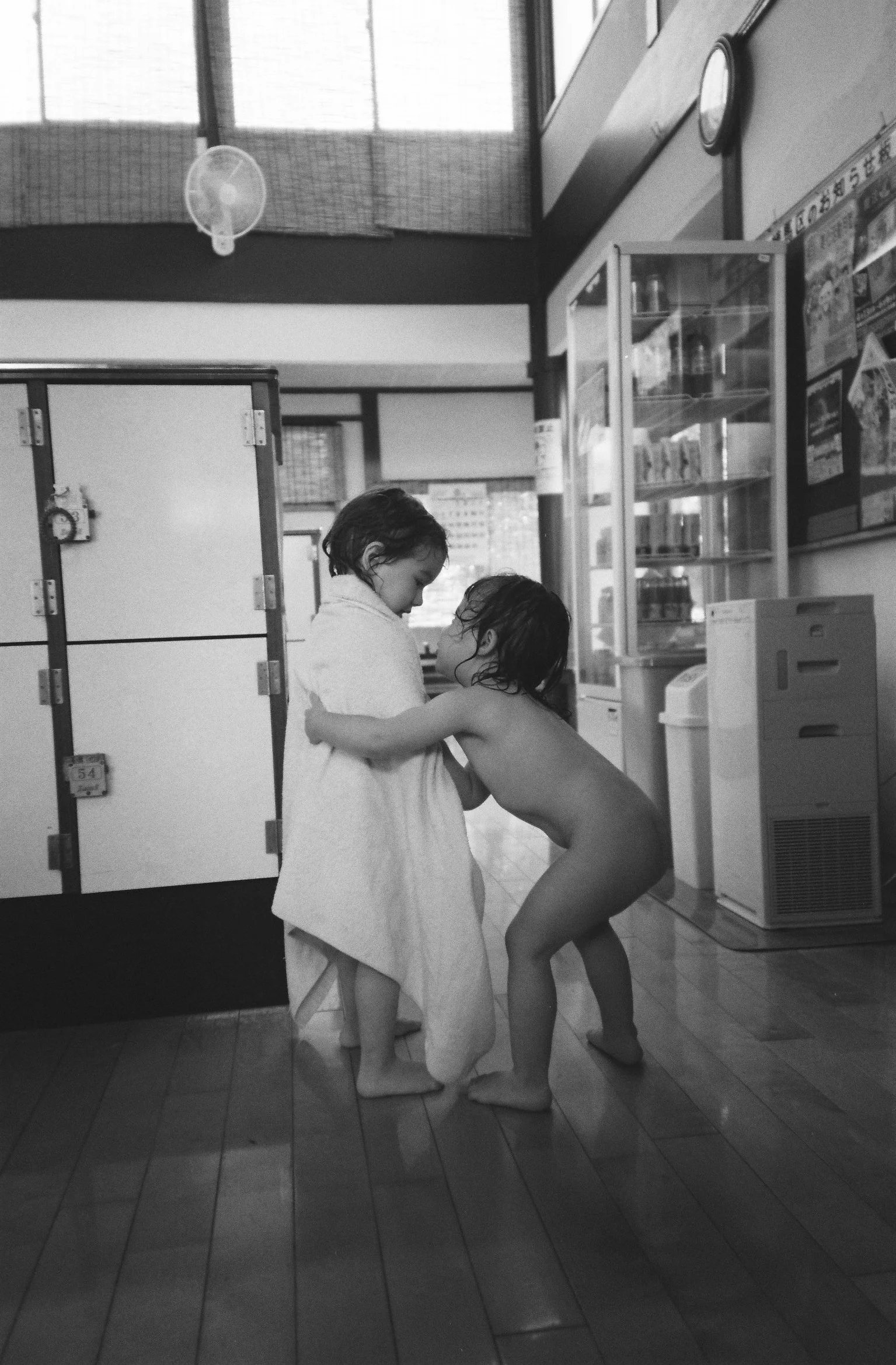 Two children after bath, one wrapping the other in a towel in a traditional public bath house Japanese sento— real family life in Japan, childhood editorial documentary captured by English speaking photographer Noa Nguyen.