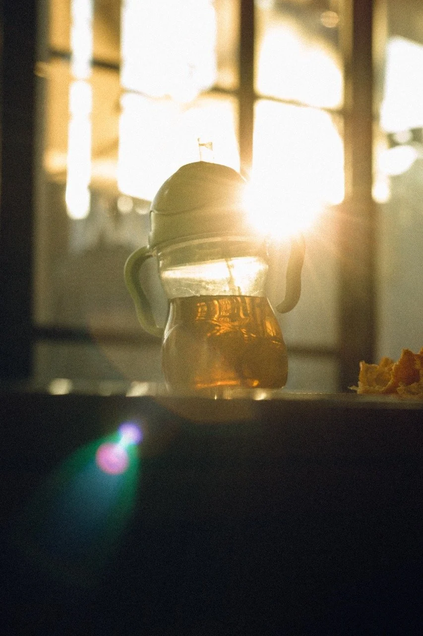 A child's sippy cup glowing in golden sunset light on a kitchen table, orange peels beside it — an ordinary Tuesday moment captured as life unfolds. Family lifestyle photography by Noa Nguyen.