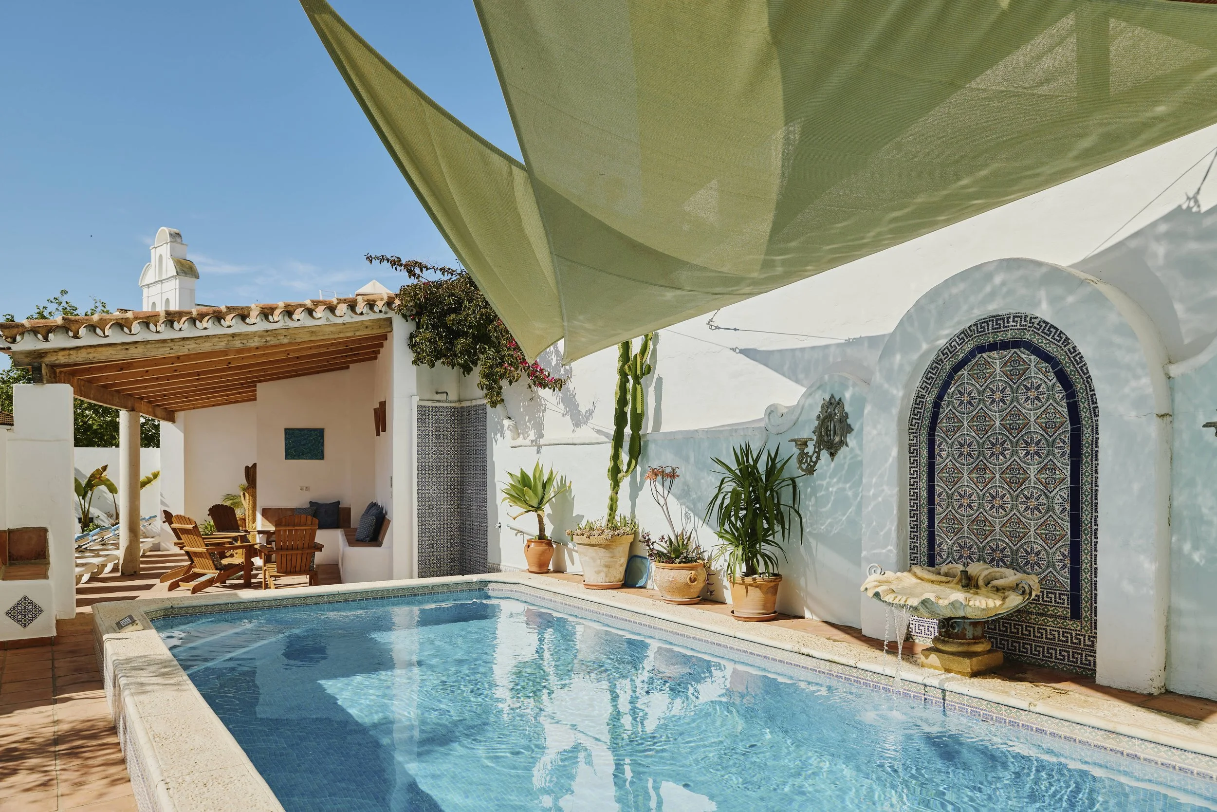 A swimming pool with white walls, potted plants, and shade sails overhead in a sunny courtyard.