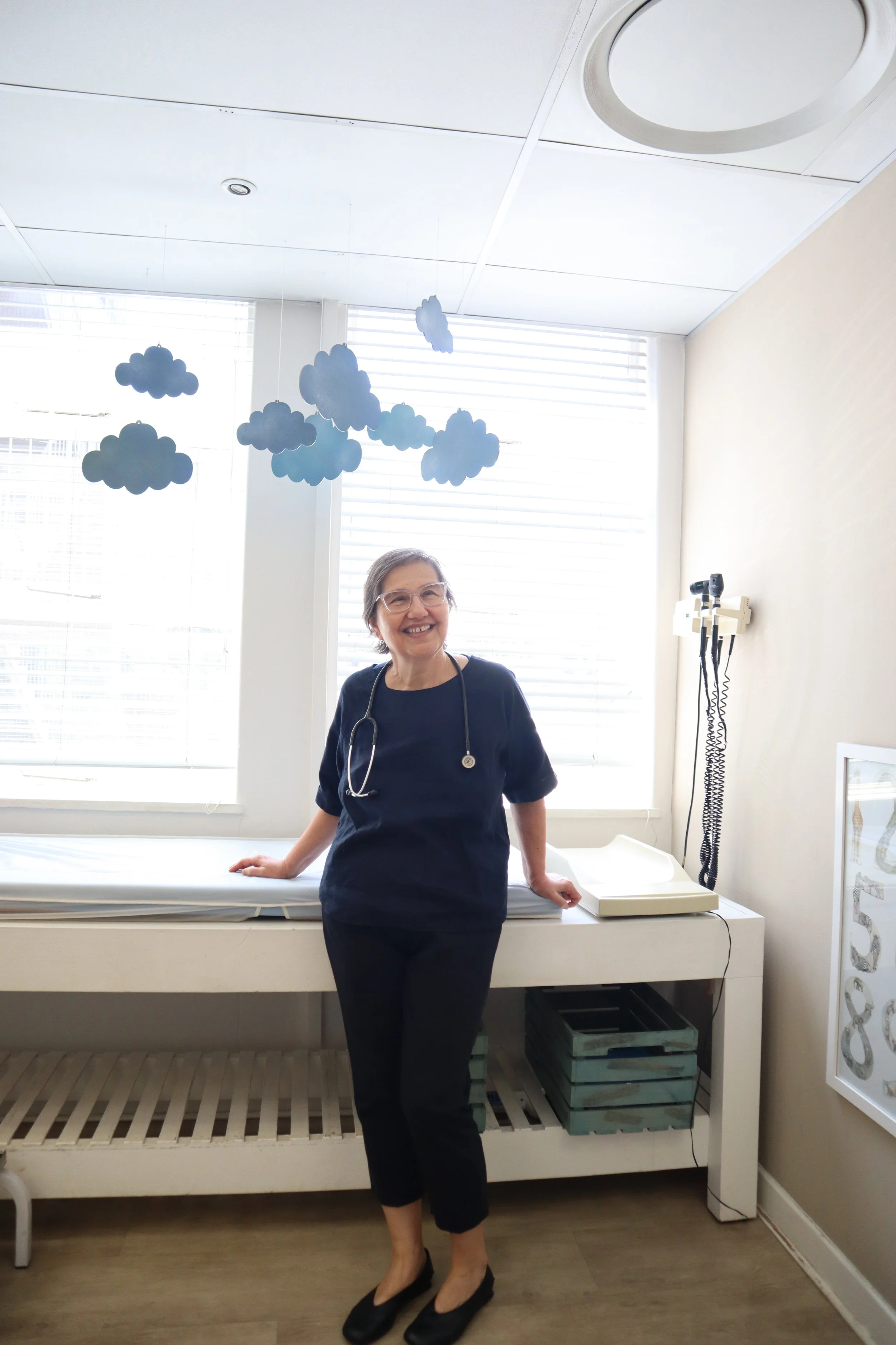 Doctor standing in medical exam room with stethoscope, smiling, near exam table and medical equipment, under decorative paper clouds.
