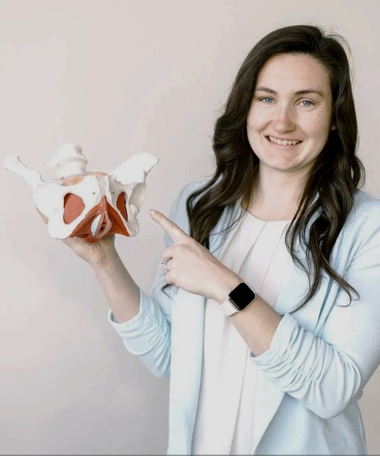 A woman smiling and pointing at a model of a human pelvis she is holding in her left hand.