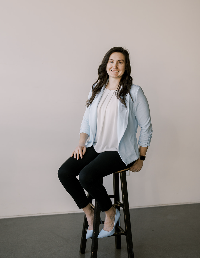 A woman sitting on a black wooden stool against a plain wall, smiling at the camera. She has long dark hair and is wearing a light blue blazer over a white blouse, black pants, and light blue high heels. She has a watch on her left wrist.