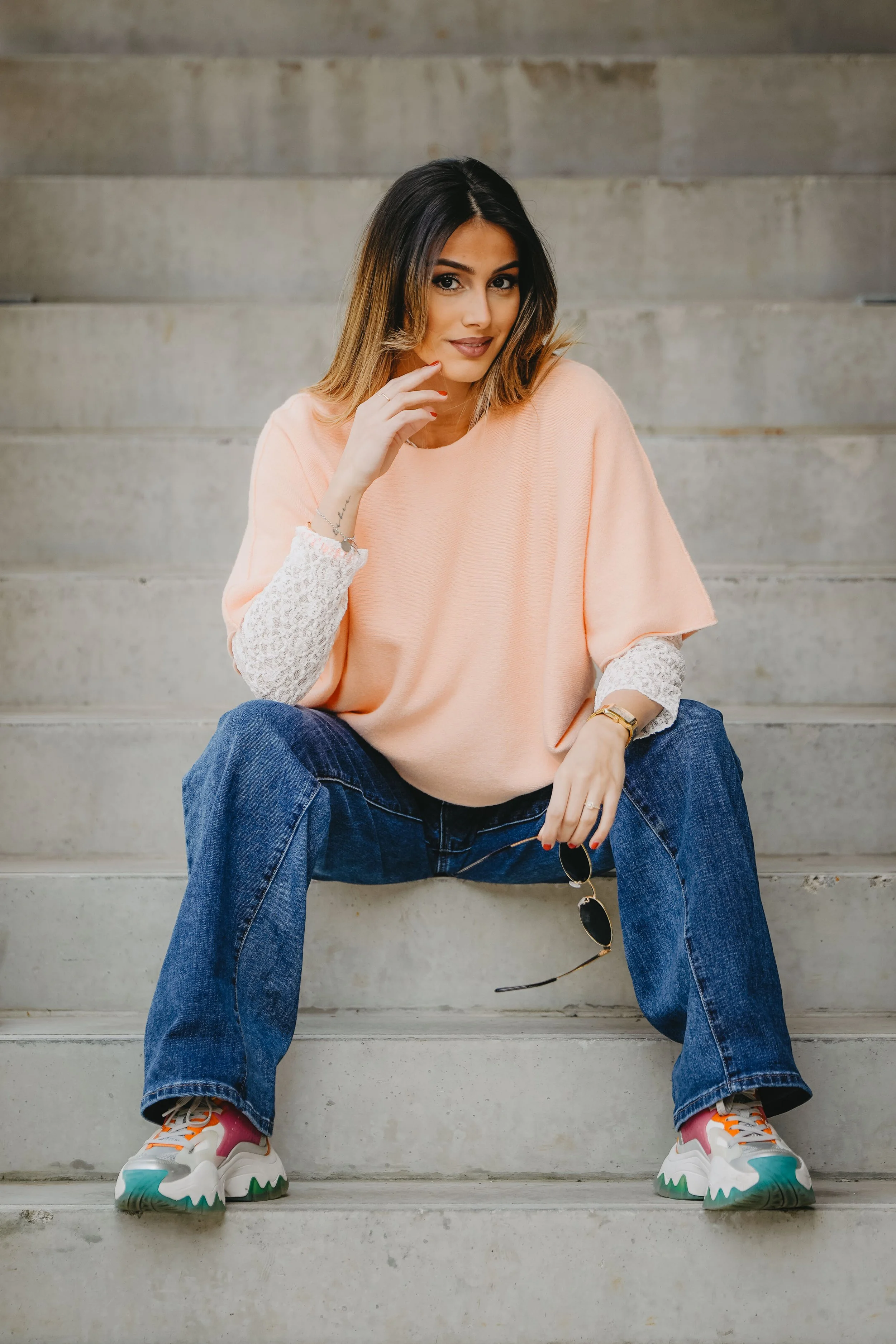 Jeune femme assise sur des escaliers en béton, portant un sweat rose, des jeans bleus, des baskets colorées, avec des lunettes de soleil dans la main.